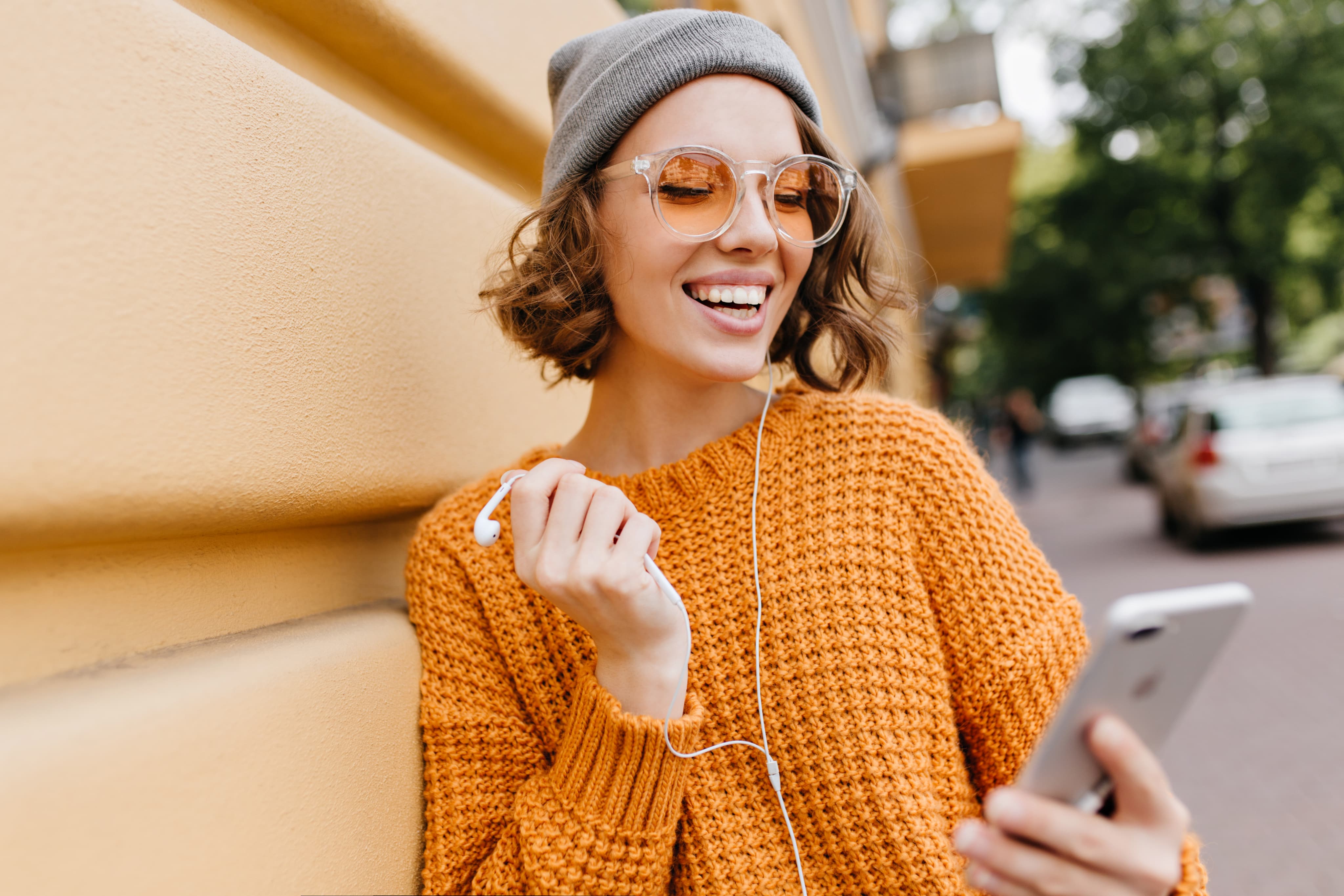 Charming short haired lady with gray hat standind beside wall with smartphone